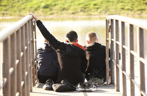 Three boys and friends are sitting on a bridge with metal handrails over the water. Friendly conversation in nature. Children's play outdoors