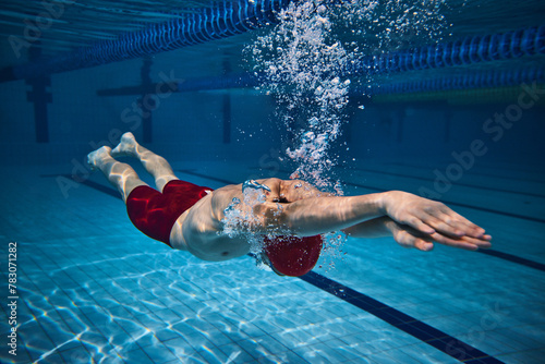 Dynamic image of athletic young guy  swimmer in cap in motion underwater  training in swimming pool. Bubbles  speed. Concept of professional sport  health  endurance  strength  active lifestyle