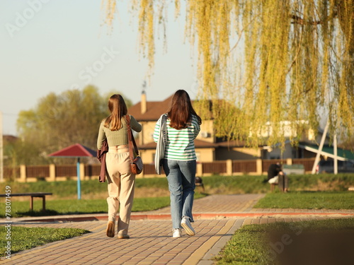 Two young girls dressed in fashionable clothes walk through a spring park in sunny weather. Girlfriends talking in nature. Walk in the fresh air.