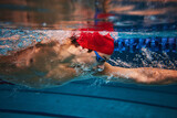Concentration. Competitive young man, swimming athlete in goggles and red cap in motion, training in pool indoors. Concept of professional sport, health, endurance, strength, active lifestyle