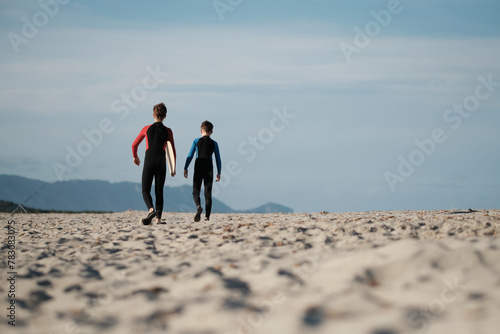 Junge Brüder im Neoprenanzug mit dem Skimboard am Strand auf Sardinien am Weg zum Wasser