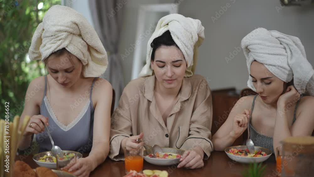 Three cute young women with towels on their heads are having breakfast while sitting at the table in the living room. Girls eat breakfast cereal with milk and look at the camera smiling Three cute young women with towels on their heads are having breakfast while sitting at the table in the living room. Girls eat breakfast cereal with milk and look at the camera smiling