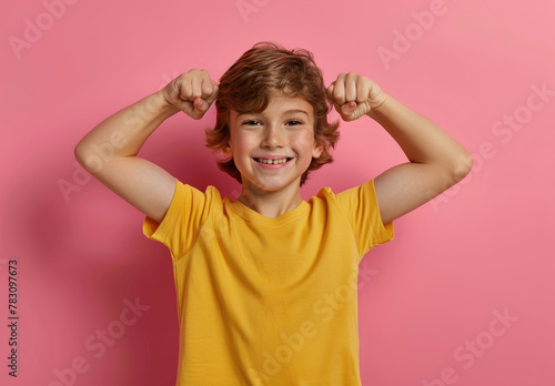 A cute little boy flexing his muscles and smiling, wearing a yellow t-shirt on a pink background