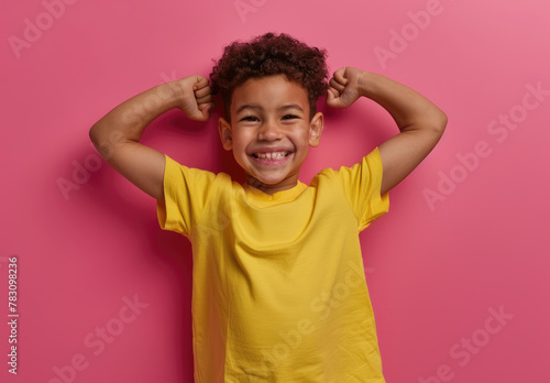 A cute little boy flexing his muscles and smiling, wearing a yellow t-shirt on a pink background