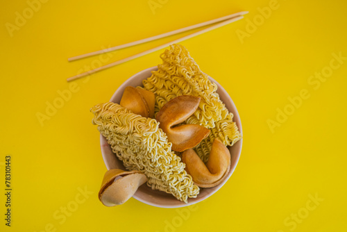A bowl with dried noodles, fortune cookies and chopsticks on yellow background. Flat lay.