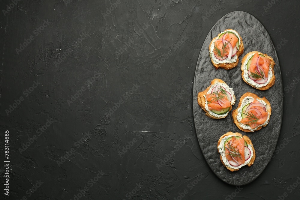 Fototapeta premium Tasty canapes with salmon, cucumber, radish and cream cheese on black table, top view. Space for text