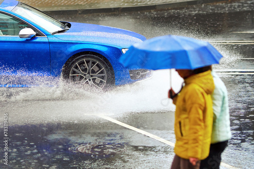 Car driving on wet road during rain, splashing water at pedestrian with umbrella. Car splashes water to pedestrian on sidewalk. Car driving on flooded street, splash water. Selective focus.
