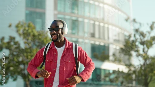 Happy cool stylish young African Black man student hipster wearing headphones, sunglasses and backpack feeling joy having fun listening music and dancing on city street outdoors on sunny day.
