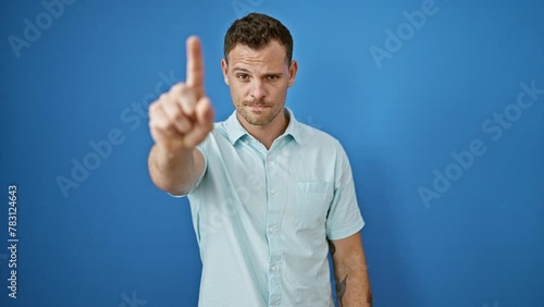 A displeased young man with beard gestures thumbs down against a blue wall background outdoor. photo