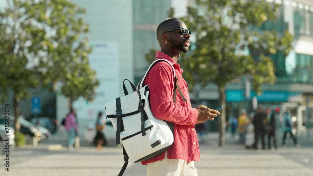 Cool young African Black man hipster student or tourist wearing sunglasses and backpack walking on city street outdoors holding smartphone using mobile cell phone technology device. Cool young African Black man hipster student or tourist wearing sunglasses and backpack walking on city street outdoors holding smartphone using mobile cell phone technology device.