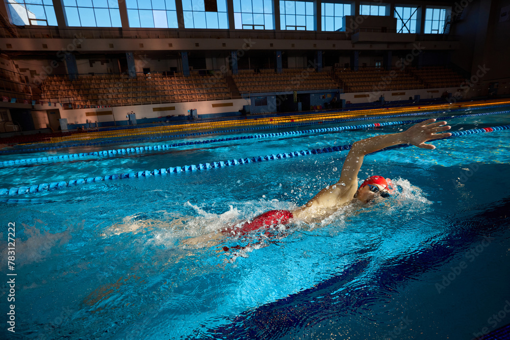 Young man, swimmer in red cap and goggles in motion, training freestyle stroke, swimming in pool indoors. Concept of professional sport, health, endurance, strength, active lifestyle