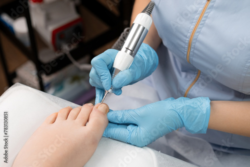 A pedicure expert in rubber gloves  performs a hardware pedicure for woman  utilizing a nail drill.