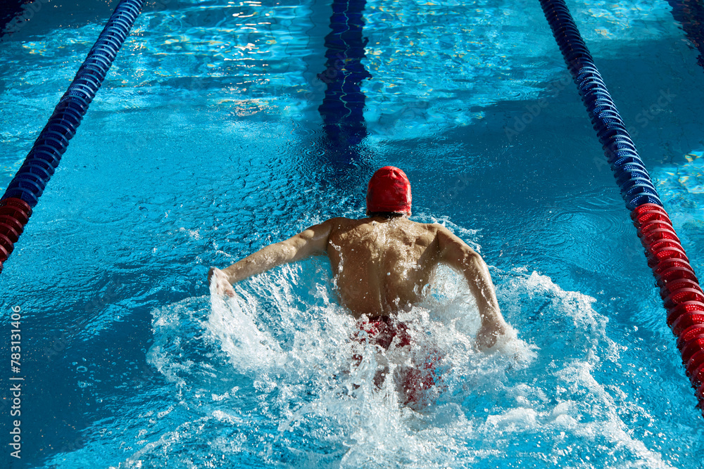 Top view of muscular, athletic young man, swimmer in red cap in motion, showing strength, training, swimming in pool indoors. Concept of professional sport, health, endurance, active lifestyle
