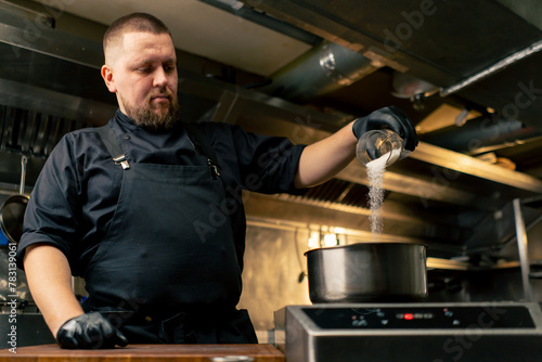 in a professional kitchen chef in a black jacket sizzles salt in a saucepan standing on the stove