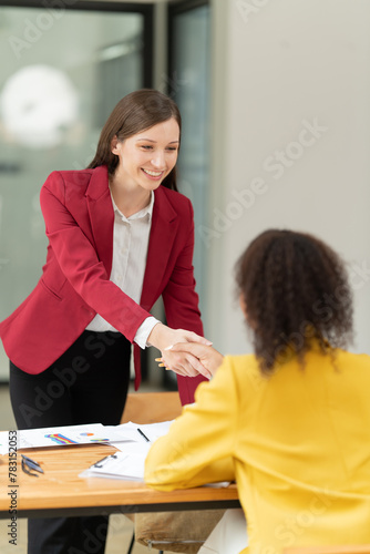 Two smiling businesswomen in professional attire shaking hands in an office setting, symbolizing a successful partnership, agreement, and warm welcome during a business meeting