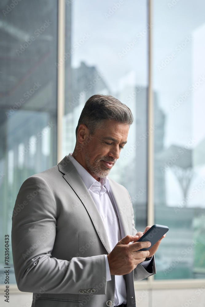 Busy elegant middle aged business man ceo, 45 year old businessman leader investor executive wearing suit holding cellphone using mobile cell phone standing in office near big window with smartphone. Busy elegant middle aged business man ceo, 45 year old businessman leader investor executive wearing suit holding cellphone using mobile cell phone standing in office near big window with smartphone.