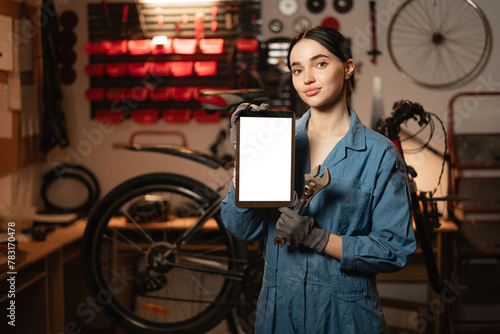 Fototapeta Naklejka Na Ścianę i Meble -  Young woman repairing bicycle in workshop, holding digital tablet with Mockup image