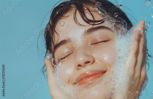 Young woman with closed eyes washing her face and smiling while using facial foam on a light blue background
