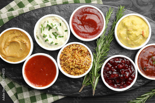 Different tasty sauces in bowls and rosemary on black wooden table, top view