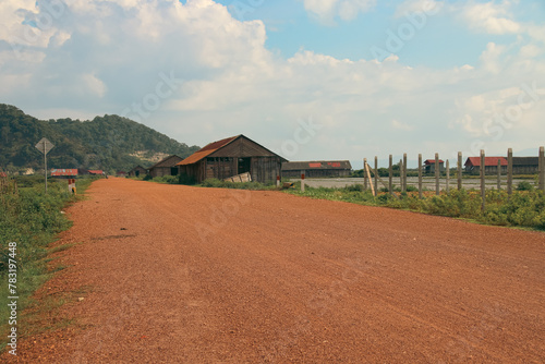 A serene rural landscape with a dirt road leading past wooden salt storage houses and mountains in the background in Fish Island, Kampot Cambodia