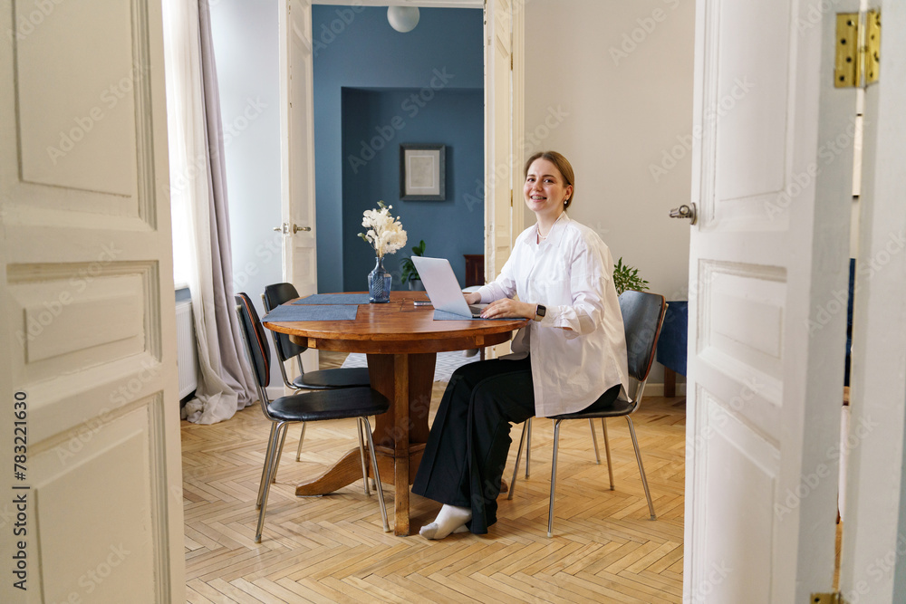 A poised woman exudes serenity at a wooden table, her laptop open, in a room awash with natural light.