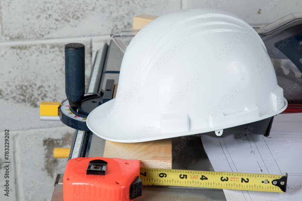 a worker's hard hat and a tape measure rest on a piece of wood lying on a table saw in a workshop
