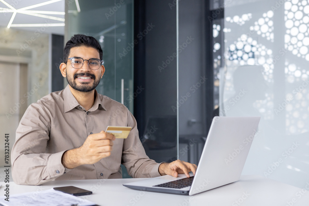 A cheerful young man in a smart shirt making an online transaction with a credit card on a laptop in a modern office setting. A cheerful young man in a smart shirt making an online transaction with a credit card on a laptop in a modern office setting.