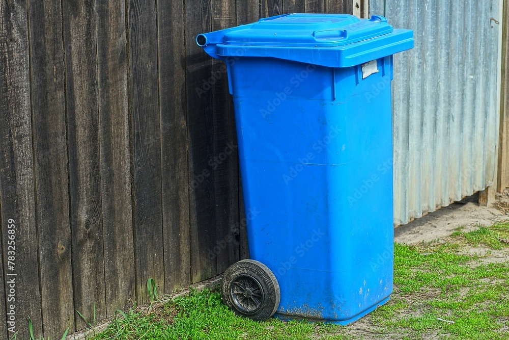 one plastic blue closed trash bin on wheels stands on the ground during the day near a wooden fence on the street one plastic blue closed trash bin on wheels stands on the ground during the day near a wooden fence on the street