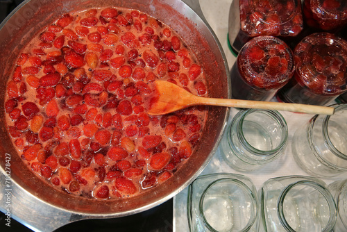 Cooking strawberry jam in a large bowl at home. Wooden spoon in a bowl with jam. Empty glass jam jars and jars filled with jam.
