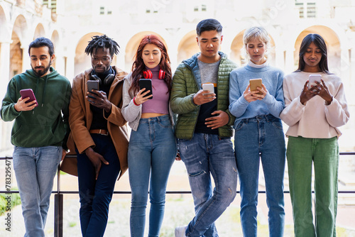 Group of young adult students stands engrossed in their smartphones - connectivity and individual isolation - social network and technology concept - university courtyard background.