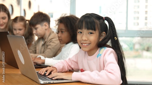 Asian girl working on laptop and looking at camera. Diverse children coding or programing engineering code or prompt by using computer in STEM technology class while teacher explain system. Erudition.