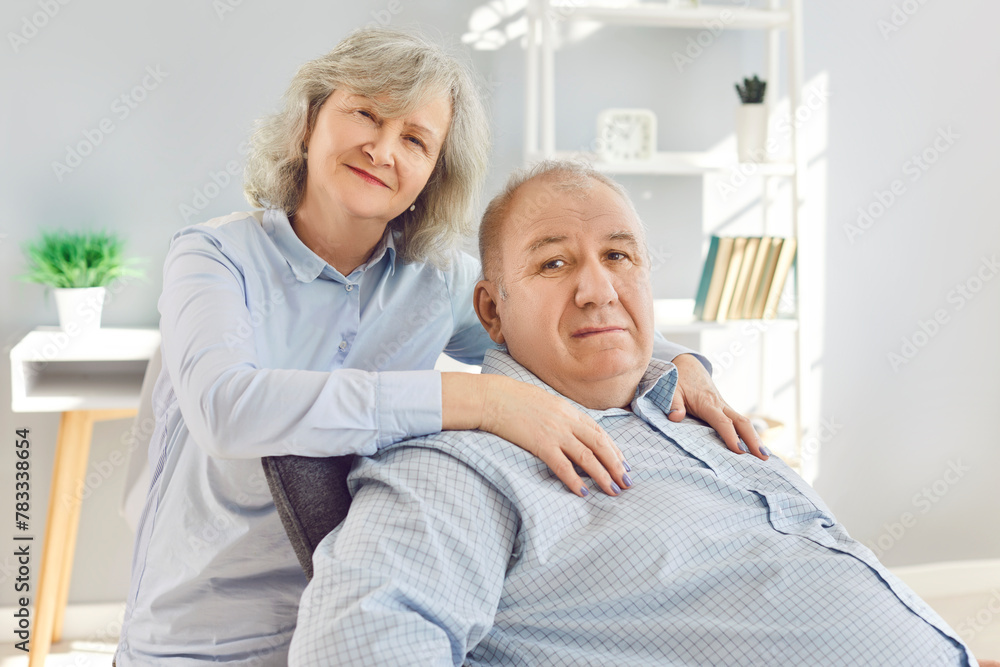 Obraz premium Portrait of a happy senior family couple wife and husband sitting in the living room at home and looking at camera. Two smiling gray-haired retired man and woman enjoying time together on retirement.