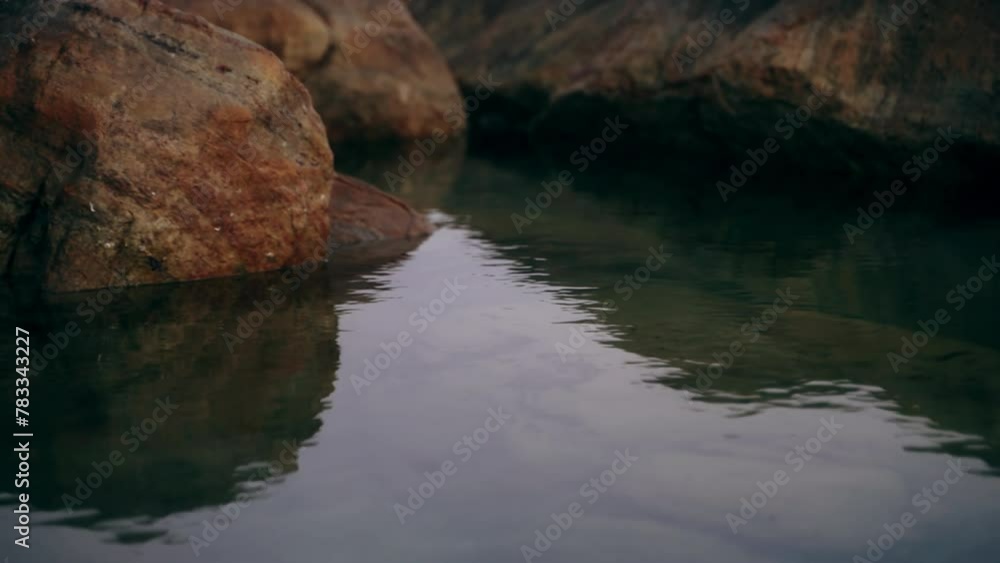 Calm sea gently laps against large weathered rocks at dusk. Reflective water at shoreline creates peaceful scene. Nature background, tranquil seaside ambiance near rocky coast in evening. Slow motion. Calm sea gently laps against large weathered rocks at dusk. Reflective water at shoreline creates peaceful scene. Nature background, tranquil seaside ambiance near rocky coast in evening. Slow motion.