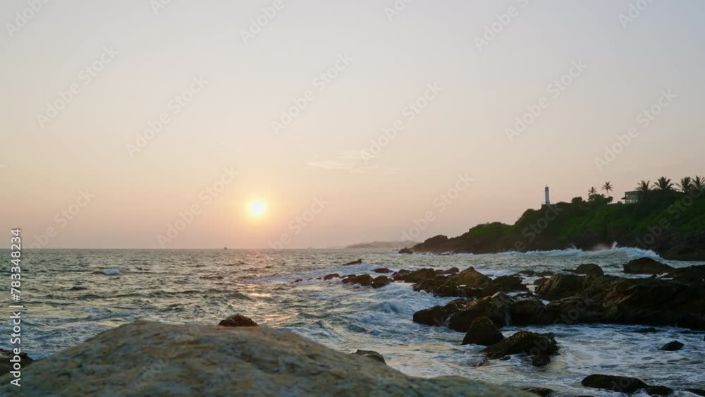 Sunset bathes lighthouse on hill in golden as waves crash against rocky shore. Serene nature, perfect for travel blog, film backdrop, eco-awareness campaign. Tranquil seascape scene invites adventure.