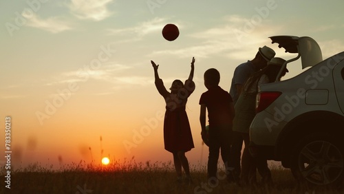 Playful family at sunset by car. cheerful children parents going on travel road trip by automobile packing baggage putting luggage in trunk kids playing ball. Packing preparation for family picnic.