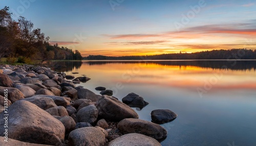 a picturesque sunset casting a golden glow over a tranquil lake with rocks scattered along the shore in the foreground under a colorful sky