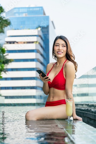 Young woman enjoys sun-kissed serenity, relaxing with drink and using phone in rooftop infinity pool