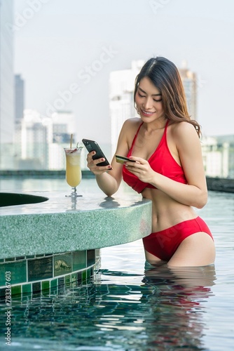 Poolside Banking: Young Asian woman in a red bikini conducts online banking on her phone while holding a credit card at the poolside bar counter