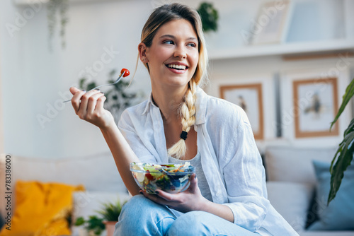 Smiling woman eating healthy salad while sitting on the kitchen at home. © nenetus