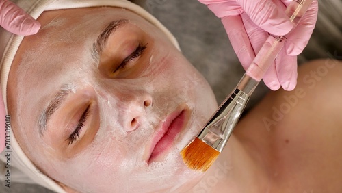 professional cosmetologist applies a white mask on the face of a female client with a brush, close-up