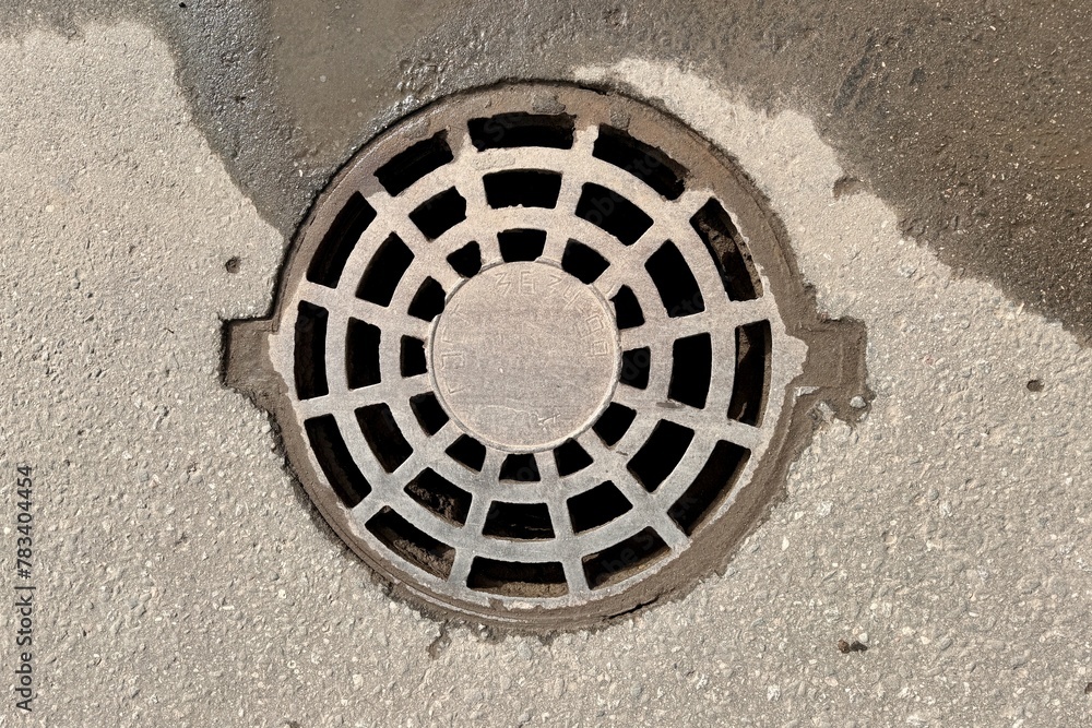 A iron hatch with a pattern above an observation well is located on auto road in daytime. Close-up
