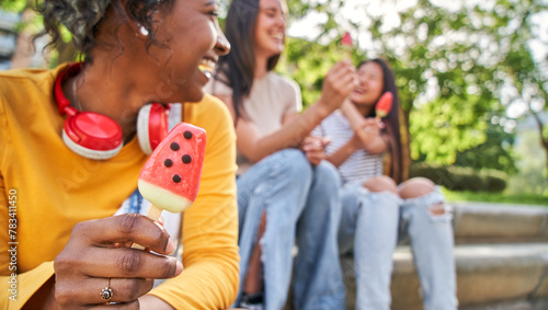 Three young multi-ethnic laughing women sitting eating ice cream in park. Female friends chatting cheerful enjoying summer afternoon together. Focus on black girl in foreground, unfocused background  photo