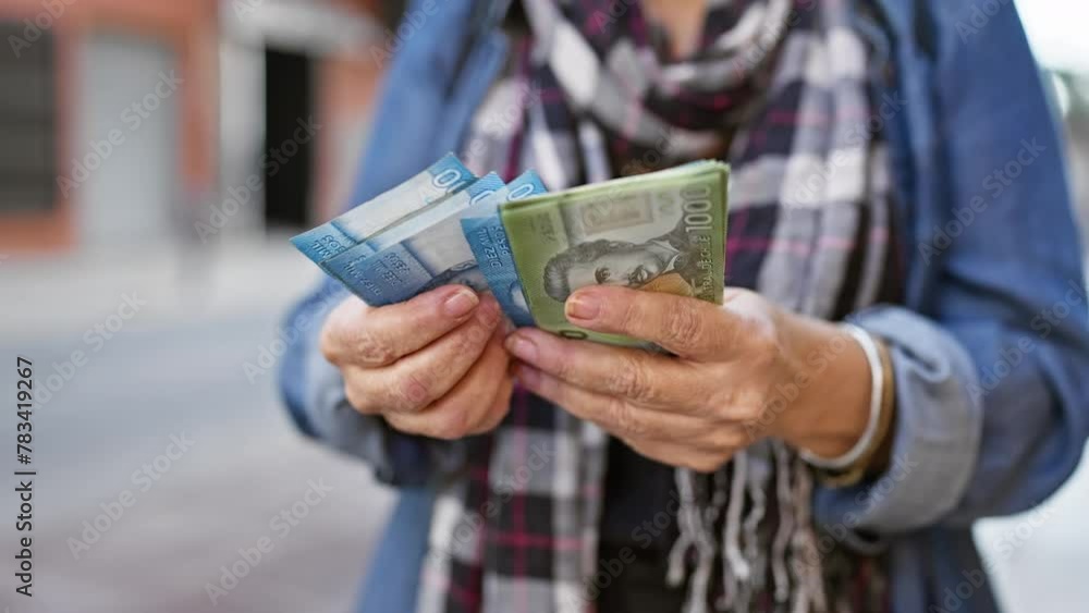 A mature woman examines chilean peso banknotes in her hands against a blurry urban street background.