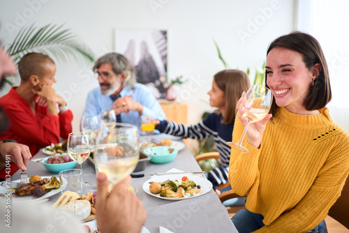 Beautiful young Caucasian woman smiling holding glass of white wine sitting at table at unfocused family meal in background. Three generations together enjoying themselves at a celebratory event