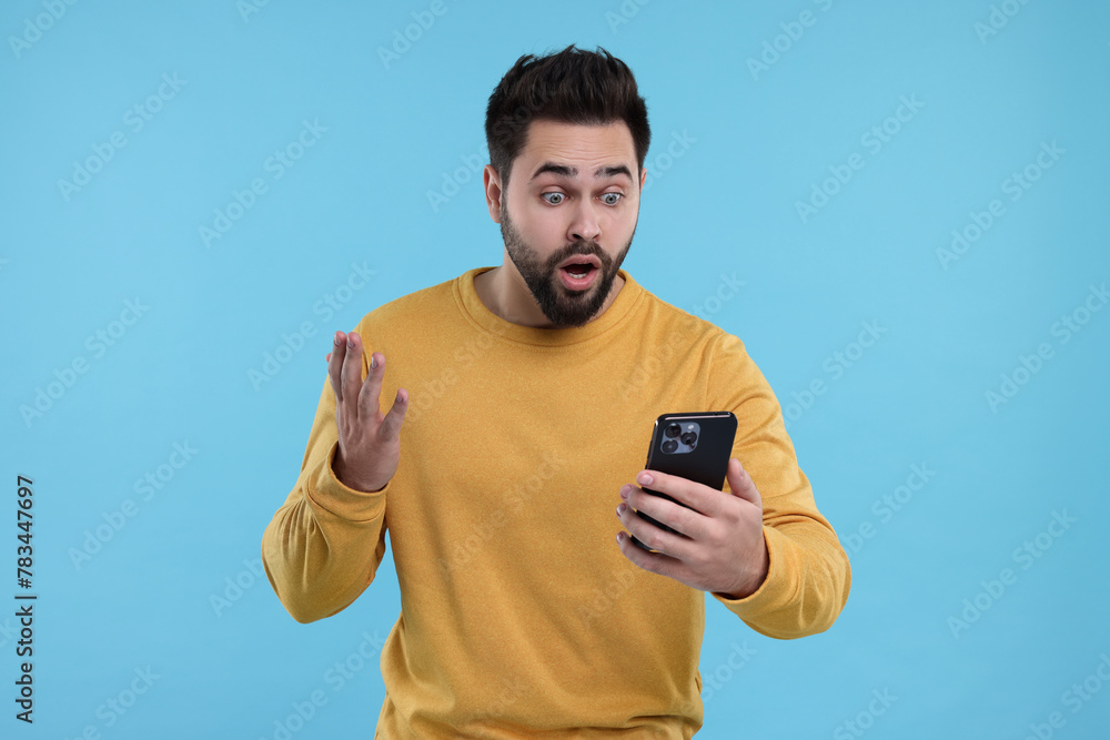 Shocked young man using smartphone on light blue background