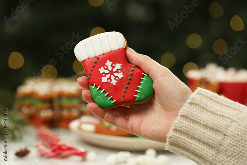 Woman with decorated cookie at table against blurred Christmas lights, closeup
