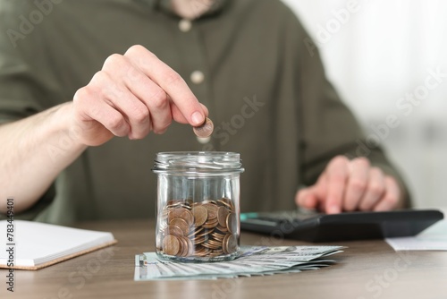 Financial savings. Man putting coin into glass jar while using calculator at wooden table, closeup