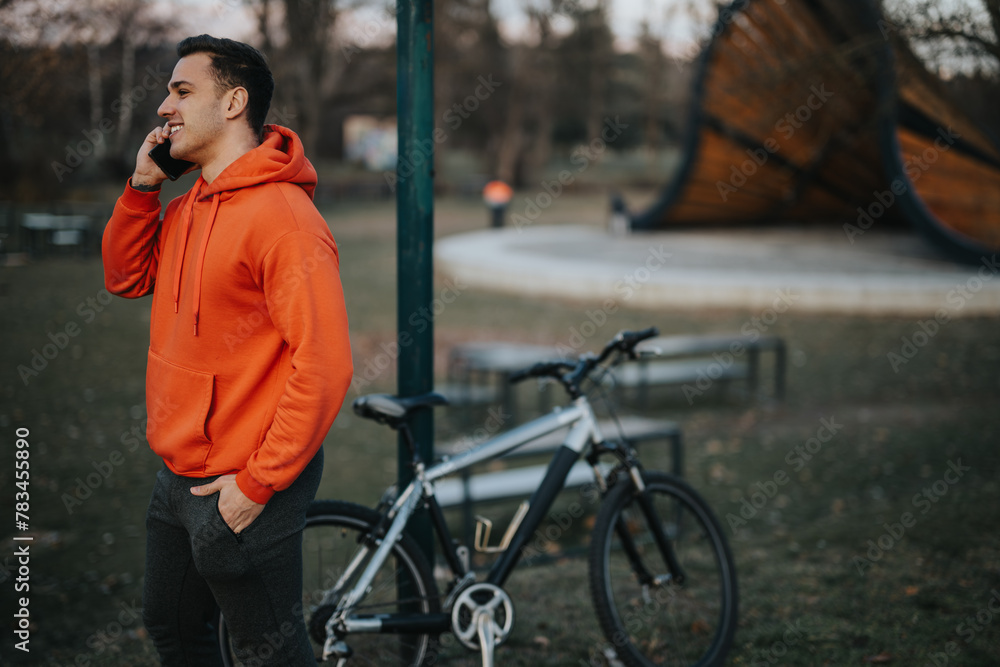 Casual young man in orange hoodie talking on phone, standing by his bicycle in an urban park setting, with the evening light. Casual young man in orange hoodie talking on phone, standing by his bicycle in an urban park setting, with the evening light.