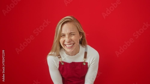 Joyful young blonde woman, wearing apron, bursts into loud, hearty laughter at hilarious joke. happily standing against isolated red background, radiating cheerful confidence.