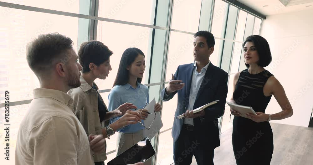 Serious Indian business team leader explaining work task to multiethnic colleagues, showing notes in notebook, speaking. Diverse team standing by large window, talking, discussing teamwork Serious Indian business team leader explaining work task to multiethnic colleagues, showing notes in notebook, speaking. Diverse team standing by large window, talking, discussing teamwork